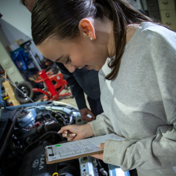 A young woman leaning over an open car engine bay, making notes on a clipboard during an automotive inspection